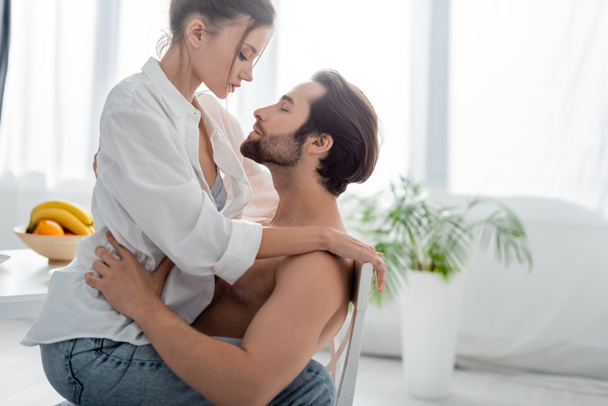 side view of seductive woman sitting on bearded and shirtless man in kitchen  - Photo, Image