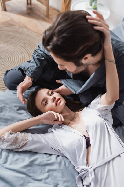 bovenaanzicht van wazig man kijken naar getatoeëerde jonge vrouw in zijden badjas liggend op bed  - Foto, afbeelding
