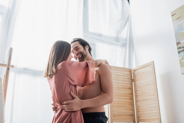 vousatý a veselý muž objímání šťastný přítelkyně v růžovém hedvábí župan - Fotografie, Obrázek