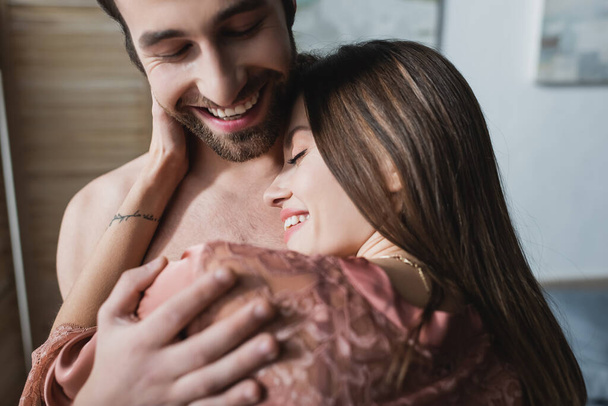 bearded and happy man hugging tattooed girlfriend in pink silk robe - Valokuva, kuva
