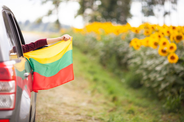 Chica sosteniendo la bandera de Lituania desde la ventana abierta del coche que conduce en una hermosa carretera. Viajar en Lituania, viajes en coche, concepto de día de la independencia. Enfoque selectivo. - Foto, imagen