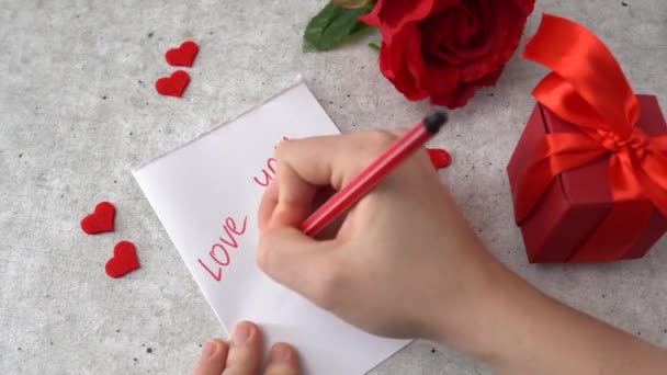 Γυναίκα ζωγραφίζει μια καρδιά για τον εορτασμό της 14ης Φεβρουαρίου. Καρδιές, τριαντάφυλλο και κόκκινο κουτί δώρου στο τραπέζι - Πλάνα, βίντεο