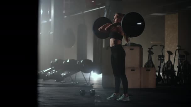 Cámara lenta: Una mujer levanta una barra en un gimnasio oscuro con mucho peso. Una joven fuerte levanta pesas para un entrenamiento - Metraje, vídeo