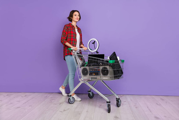 Full length photo of cute young brunette lady go with conservation trolley wear shirt jeans shoes isolated concrete violet wall background - Fotoğraf, Görsel