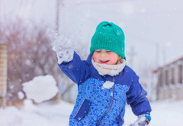 cute child boy in a knitted green hat and white scarf on a snowy street playing snowballs. winter holidays, outdoor activities in the snow, childhood. - Photo, Image