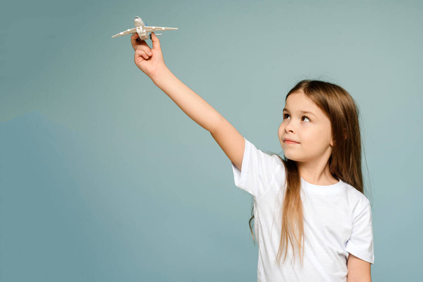 Φωτογραφία ενός μικρού ευτυχισμένου κοριτσιού που κρατάει ένα αεροπλάνο-παιχνίδι. Αντιγραφή απομονωμένου χώρου σε φόντο μπλε χρώματος - Φωτογραφία, εικόνα
