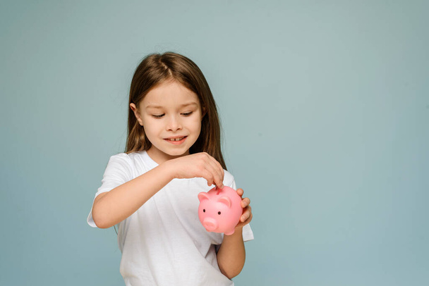 A little girl puts a coin in a piggy bank. The concept of teaching children personal finance and saving. - Zdjęcie, obraz