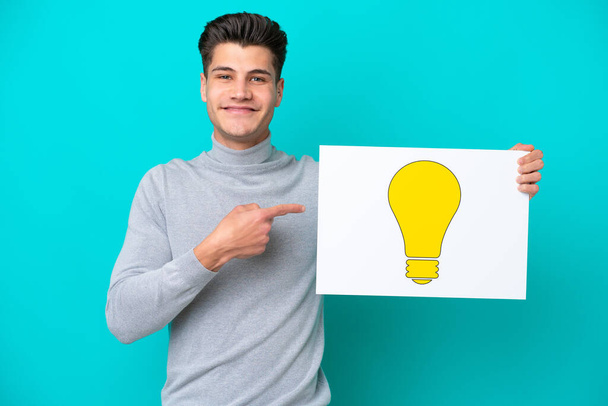 Junger gutaussehender kaukasischer Mann, isoliert auf blauem Grund, hält ein Plakat mit einem Glühbirnensymbol in der Hand und zeigt darauf - Foto, Bild