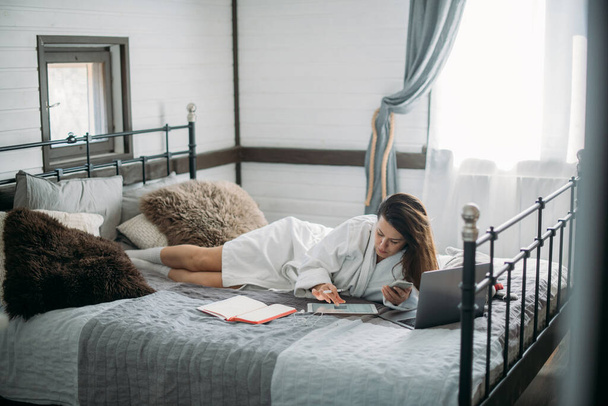 Mulher trabalha com laptop e tablet na cama no quarto. Uma jovem deita-se em uma cama grande em casa, trabalha online, toma notas em um tablet, se comunica com os clientes
 - Foto, Imagem
