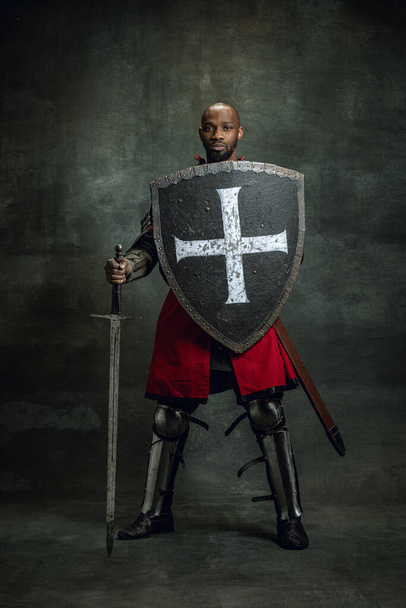 Portrait of medieval warrior, dark skinned bald knight with dirty wounded face holding shield and sword isolated over dark vintage background. Comparison of eras, history, art - Fotografie, Obrázek