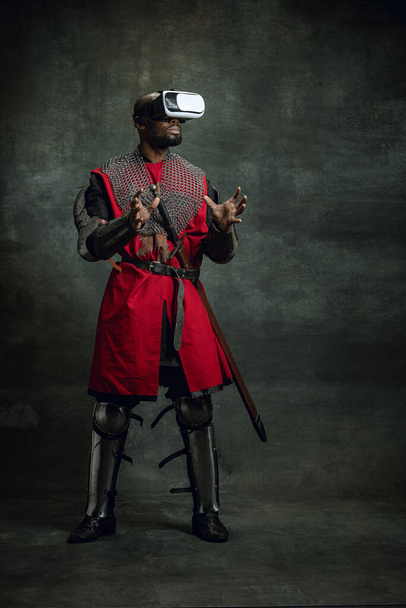 Moderne Gadgets. Ein dunkelhäutiger Mann, mittelalterlicher Krieger oder Ritter mit VR-Headsets vor dunklem Vintage-Hintergrund. Vergleich von Epochen, Geschichte, Technologie - Foto, Bild