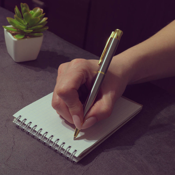 A woman's hand holds a silver pen and writes in a notebook on the table. The concept of Business, freelancing, planning, tonning - Foto, afbeelding