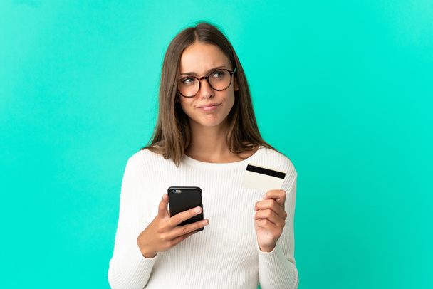 Mujer joven sobre aislado fondo azul compra con el móvil con una tarjeta de crédito, mientras que pensando - Foto, imagen