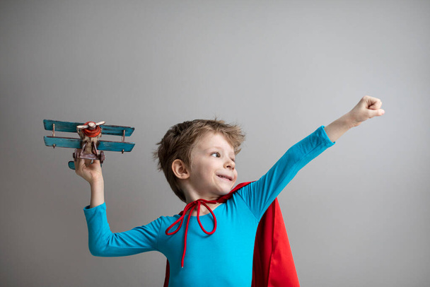 Little superhero boy in a red cape plays with an airplane. Happy smiling child. Success, motivation and travel concept. - Photo, image