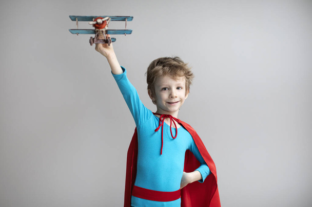 Little superhero boy in a red cape plays with an airplane. Happy smiling child. Success, motivation and travel concept. - Foto, Imagen