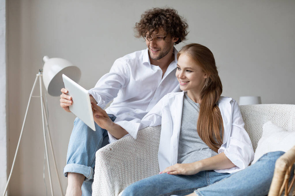 Young beautiful couple in cozy wicker chair with digital tablet online shopping, wear jeans and white shirts - Foto, Imagem