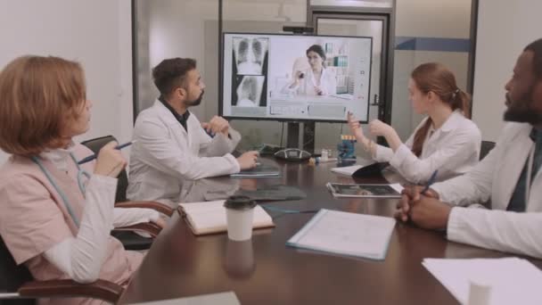 Dollying-out-Aufnahme einer Gruppe verschiedener Ärzte, die ein Videotreffen mit einer Kollegin auf dem Bildschirm haben und über das Röntgen der Brust einer Patientin diskutieren, die im Konferenzraum am Tisch sitzt - Filmmaterial, Video