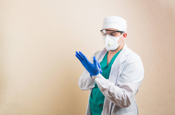 Young doctor in white suit and protective breathing mask puts second gloves on his hands to protect his hands from dangerous disiase. Isolated on yellow background. - Photo, Image