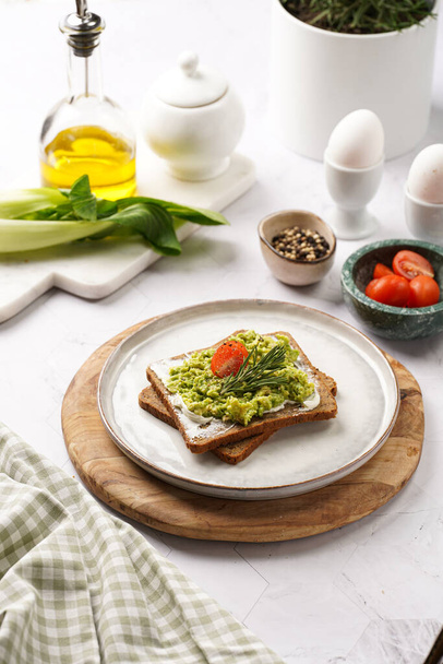 Trendy lifestyle sandwich: protein bread slice with cream cheese, mashed avocado, cherry tomatoes and rosemary on white scandi plate, light setting - Фото, изображение