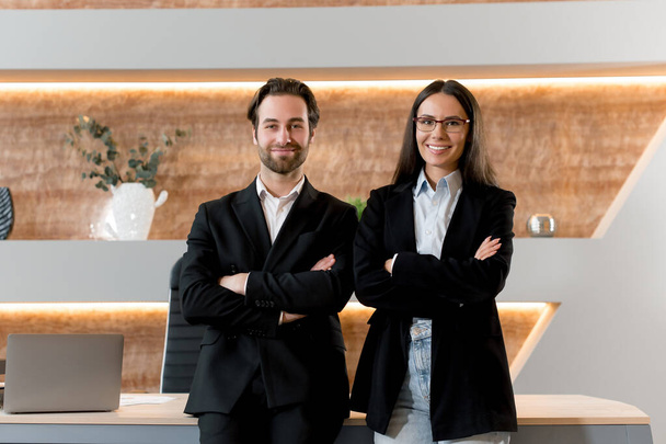 Successful intelligent caucasian young adult man and woman, office colleagues, financials, entrepreneurs, standing at the desktop, in formal and stylish clothes, arms crossed, friendly smiling - Фото, изображение