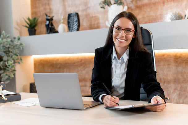 Portrait of a successful, young adult business woman, broker, financial consultant, sales manager, sit in a modern office, in stylish business clothes, working with papers, looks at camera, smiling - 写真・画像