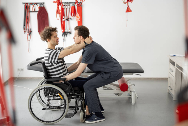 Rehabilitasyon uzmanı, klinikteki tekerlekli sandalyeden kalkmasına yardım ediyor. - Fotoğraf, Görsel