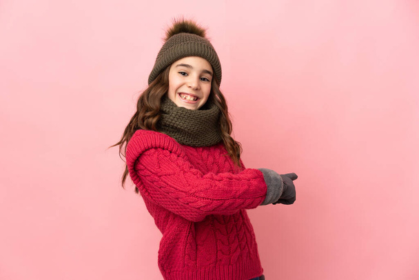 Little girl with winter hat isolated on pink background pointing back - Фото, изображение
