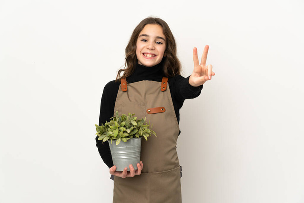 Petite fille tenant une plante isolée sur fond blanc souriant et montrant un signe de victoire - Photo, image