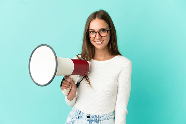 Young woman over isolated blue background holding a megaphone and smiling a lot - Fotoğraf, Görsel