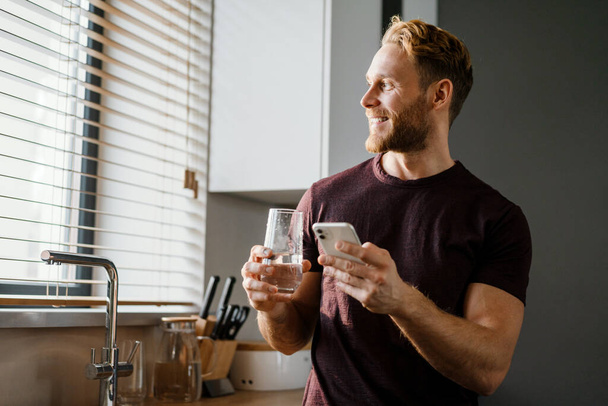 Attractive young man texting on his smartphone while drinking water from a glass at home - Foto, Imagem