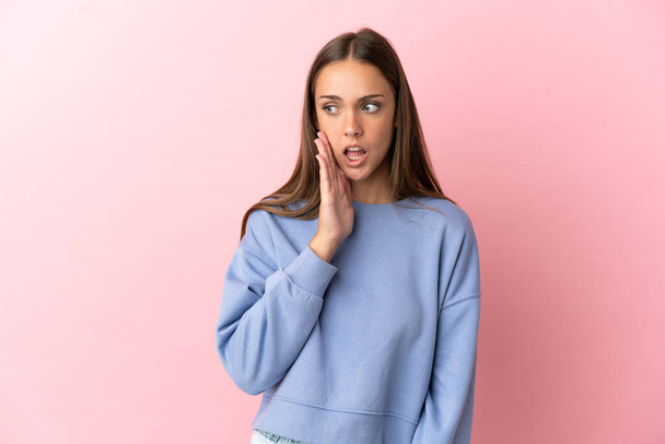 Young woman over isolated pink background whispering something with surprise gesture while looking to the side - Photo, Image
