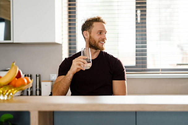Smiling healthy man drinking water from a glass while standing in the kitchen - 写真・画像