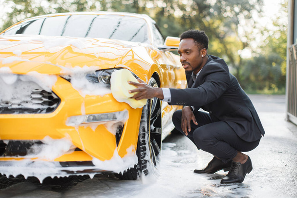 African man in business suit, washing his yellow car headlights with yellow sponge and soap. - Photo, image