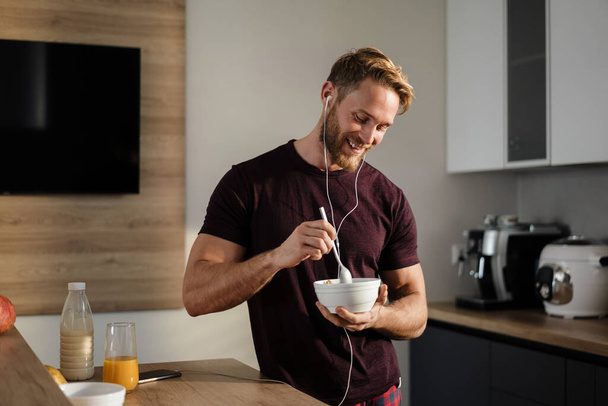 Jeune homme sain attrayant prenant un délicieux petit déjeuner tout en restant debout dans la cuisine, tenant bol, écoutant de la musique avec des écouteurs - Photo, image