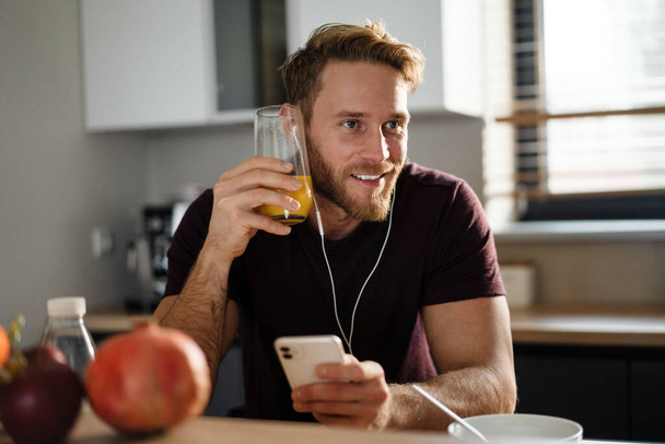 Handsome young man listening to music with earphones, browsing on mobile phone while sitting in the kitchen, eating apple - Photo, image