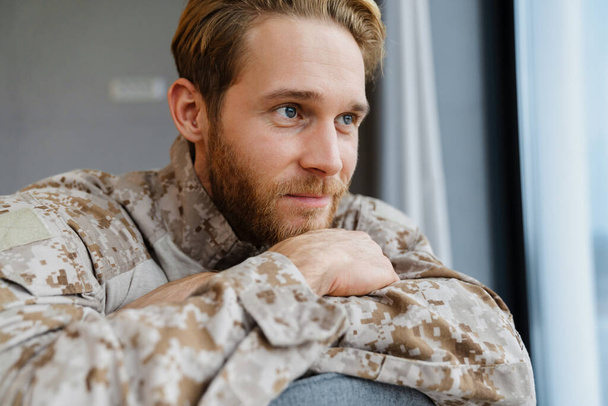 Masculine pleased military man looking aside while sitting on chair indoors - Foto, Imagen