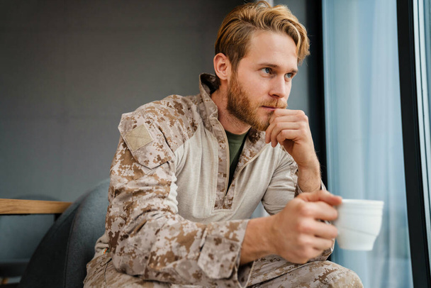 Męski pewny siebie wojskowy w mundurze siedzi i pije kawę w domu - Zdjęcie, obraz