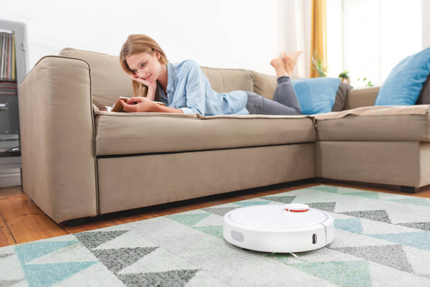 Modern robotic vacuum cleaner. Autonomous robot cleans the apartment, young woman has free time - 写真・画像