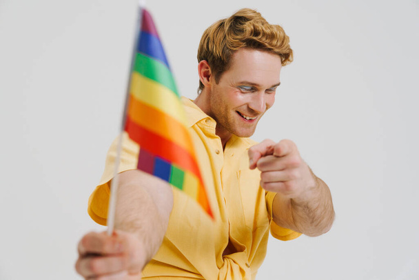 Ginger young man showing rainbow flag and pointing finger at camera isolated over white wall - Photo, image