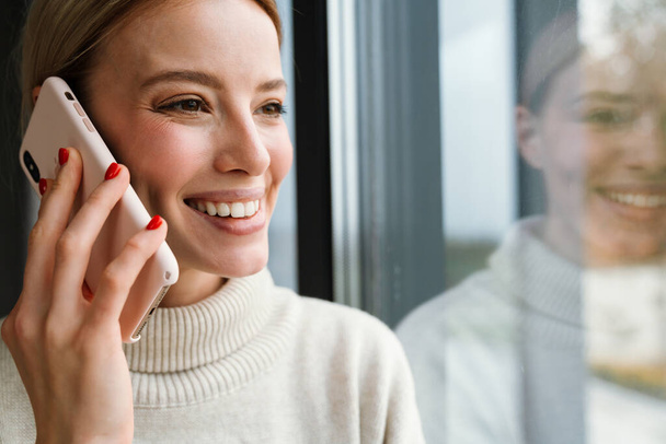Beautiful smiling woman talking on mobile phone while leaning on window glass indoors - Foto, Imagem