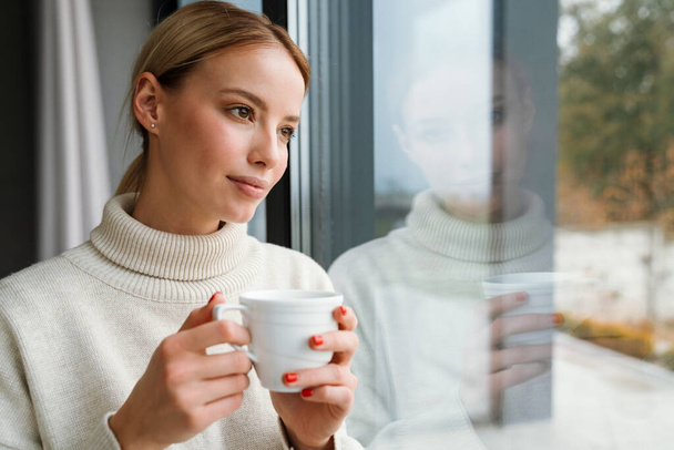 Beautiful pleased woman drinking coffee while leaning on window glass indoors - Photo, Image