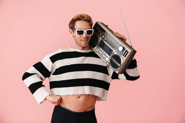 Ginger young man wearing sunglasses posing with retro tape recorder isolated over pink wall - Fotó, kép