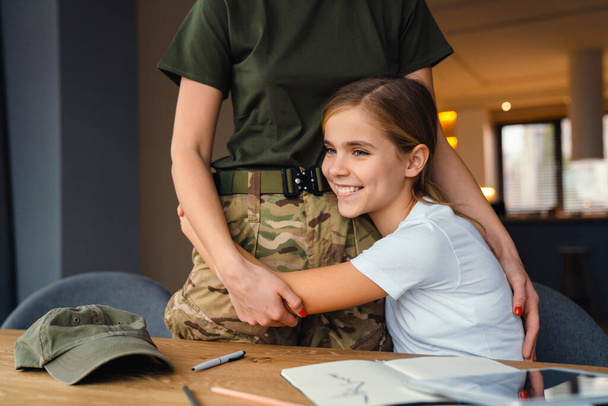 Bella donna militare che si abbraccia mentre fa i compiti con sua figlia a casa - Foto, immagini