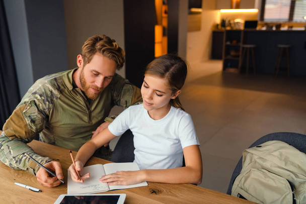 Masculine pleased military man doing homework with her daughter at home - Foto, Imagen