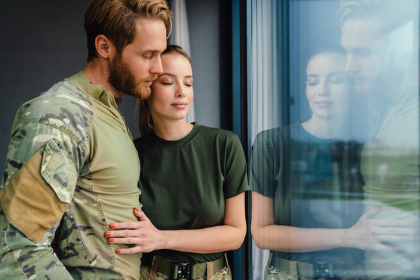Красивая молодая военная пара обнимается и смотрит в окно в помещении - Фото, изображение