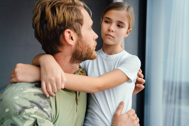 Masculine military man hugging her daughter while kneeling indoors - Zdjęcie, obraz