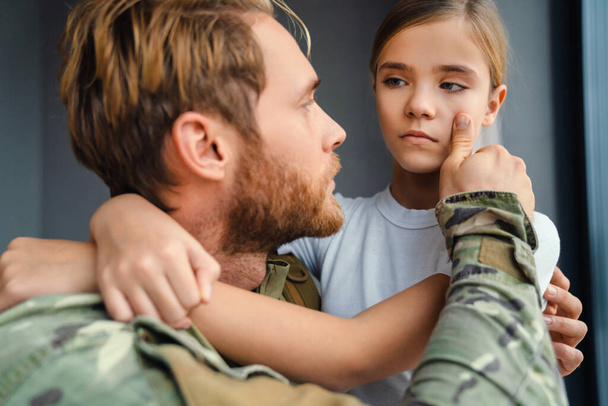 Мужчина-военный обнимает ее плачущую дочь, стоя на коленях в помещении - Фото, изображение