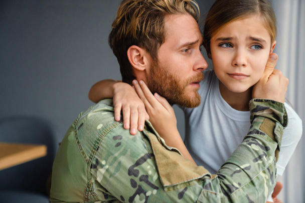 Masculine military man hugging her crying daughter while kneeling indoors - Photo, image