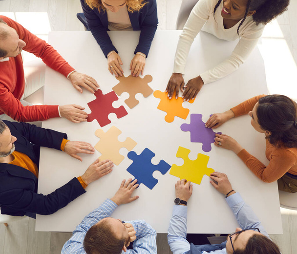 Business team sitting around office table and putting jigsaw puzzle pieces together - Фото, изображение