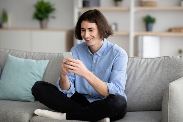 Uusi mobiilisovellus. Onnellinen nuori kaveri, joka käyttää älypuhelinta, kutoma ja selailu internetissä istuessaan rajat jalkainen sohvalla - Valokuva, kuva